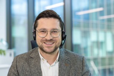 Genç bir ofis çalışanının yakın plan portresi. İş yerinde kulaklık ve takım elbiseyle oturuyor ve gülümseyerek kameraya bakıyor..