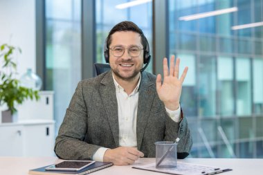Ofisteki bir masada oturan, gülümseyen ve kameraya el sallayan takım elbiseli bir iş adamının portresi..