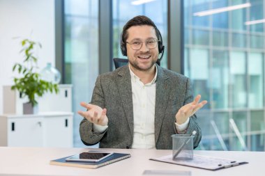 Ofisteki bir masada oturan, takım elbiseli ve kulaklıklı, kameraya açıklama yapan ve konuşan genç bir adamın portresi..