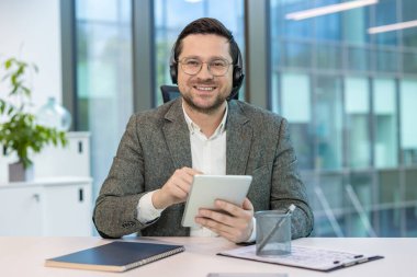 Kulaklık ve gözlüklü genç bir adamın portresi çalışma masasında kameranın önünde oturuyor, elinde bir tablet tutuyor ve gülümseyerek kameraya bakıyor..