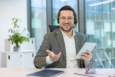 Kulaklık ve takım elbiseli gülümseyen başarılı bir iş adamının portresi bir masada oturmuş elinde tabletle kamerayla konuşuyor..
