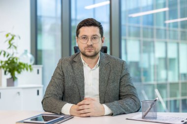 Gözlüklü, takım elbiseli, genç ve ciddi bir işadamının yakın plan portresi. Masasında oturmuş kameraya bakıyor..