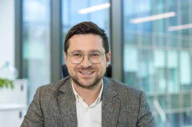 Ofisinde oturan ve kameraya bakan gözlüklü ve takım elbiseli genç bir işadamının portresi..