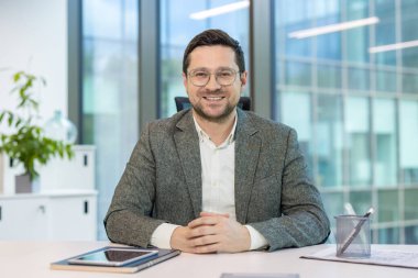 Gülümseyen ve kendinden emin bir ofis çalışanı ve iş adamının portresi masasında oturmuş kameraya bakıyor..