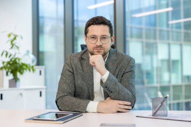 Ciddi ve başarılı bir şirket CEO 'su ve yönetmeni bir ofiste takım elbiseli bir şekilde oturur, elini yüzüne yaklaştırır ve kameraya güvenle bakar..