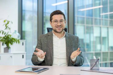 Gülümseyen genç erkek işadamı masada oturuyor ve video görüşmesiyle çevrimiçi bir konferans düzenliyor, elleriyle kameraya işaret ediyor..