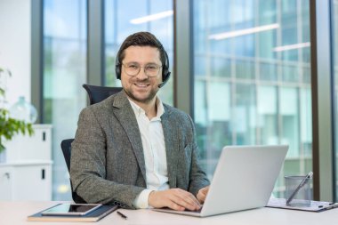 Takım elbiseli ve kulaklıklı gülümseyen genç bir adamın portresi çalışma masasında oturup kameraya bakıyor..