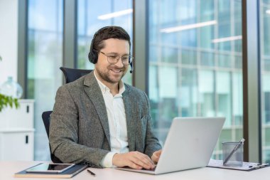 Gülümseyen, kulaklıklı genç bir ofis çalışanı ofiste oturuyor ve dizüstü bilgisayarla çalışıyor..