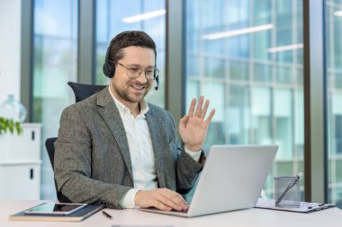 Gülümseyen genç bir ofis çalışanı masasında bir kulaklıkla oturuyor ve dizüstü bilgisayarının kamerasına el sallıyor..