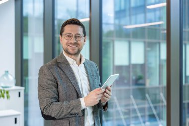 Ofiste elinde tabletle kameraya bakan takım elbiseli ve gözlüklü başarılı ve gülümseyen genç bir işadamının portresi..