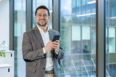 Ofiste duran takım elbiseli ve gözlüklü gülümseyen genç bir iş adamı cep telefonunu tutuyor ve kameraya gülümsüyor.