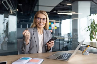 Bir iş kadını ofis ortamında elinde bir telefon ve dizüstü bilgisayarla masasında başarıyı kutluyor..