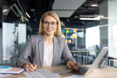 Modern ofis ortamında gülümseyen bir iş kadını, not alırken dizüstü bilgisayarda çalışıyor, kendine güvenini yayıyor..