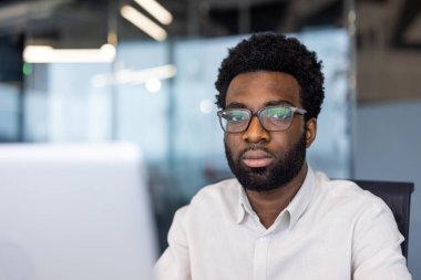 Gözlüklü ciddi bir adam ofis ortamında kameraya bakar..