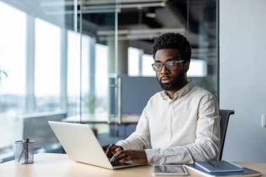 Gözlüklü bir adam modern ofis ortamında dizüstü bilgisayarda çalışıyor. Kendine güvenen ve profesyonel görünüyor..