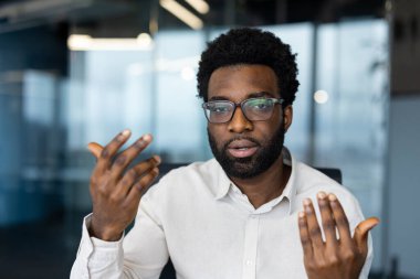 Video konferansı sırasında gözlüklü bir adam. Bir şey konuşuyor gibi görünüyor, eller hareket ediyor..