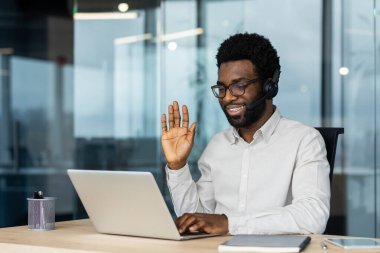 Videoda gülümseyen bir adam, kulaklık ve gözlük takıyor, ofis ortamında dizüstü bilgisayara el sallıyor..