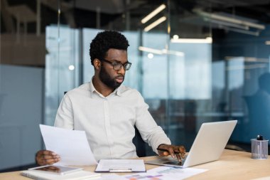Gözlük takan Afrikalı bir iş adamı modern ofis ortamında dizüstü bilgisayarda çalışıyor, belgeleri inceliyor..