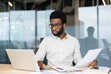 Gözlüklü Afrikalı Amerikalı bir adam ofiste bir masada oturuyor, bir dizüstü bilgisayar üzerinde çalışıyor ve belgeleri inceliyor..