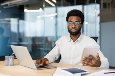 Elinde tablet tutarken bilgisayarla çalışan odaklanmış Afro-Amerikalı bir adam, bir ofisteki kameraya bakıyor..