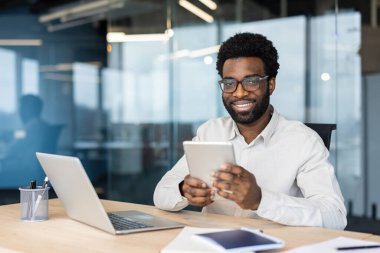 Gözlüklü, gülümseyen bir Afro-Amerikalı ofis masasında tablet kullanıyor..
