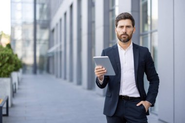 Kendine güvenen bir işadamı elinde bir tabletle dışarıda duruyor. Takım elbise giymiş, odaklanmış bir ifadeyle modern teknolojiyi sergiliyor..
