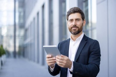 Takım elbiseli bir işadamı bir ofis binasının önünde dururken tablet tutuyor. Doğrudan kameraya bakıyor, kendinden emin..