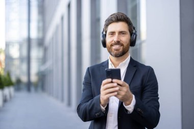 Kulaklıklı takım elbiseli bir işadamı, şehirde akıllı telefon tutarken gülümsüyor..