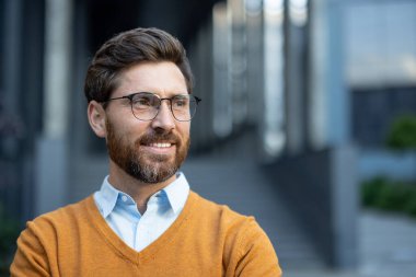 Sakallı, gözlüklü ve hardal kazaklı gülümseyen bir adam yan tarafa bakıyor..