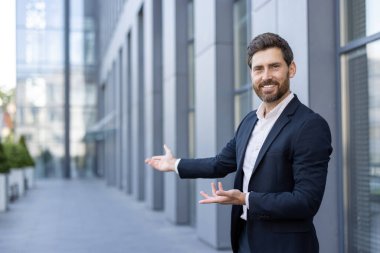 Takım elbiseli bir işadamı iki eliyle modern bir binanın önünde bir şeyi hoş karşılar ya da sunar..