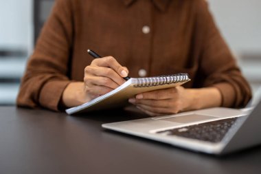 Bir kişi bir spiral deftere kalemle yazar, arka planda görünen bir dizüstü bilgisayarla, verimliliği yakalar ve not alır..