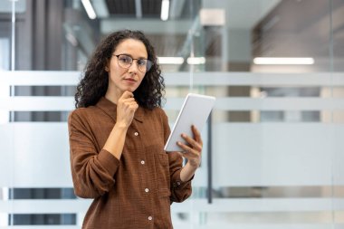 Gözlüklü düşünceli bir kadın, elinde tablet, içeride cam duvarlı bir şey düşünüyor..