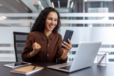 Ofisteki bir kadın gülümsüyor, elinde telefonla kutlama yapıyor. Masada bir dizüstü bilgisayar, tablet ve defter var..