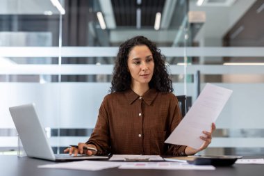 Bir iş kadını modern bir ofiste çalışır, bir dizüstü bilgisayar kullanırken belgeleri analiz eder, üretkenliği ve profesyonel odaklanmayı vurgular.