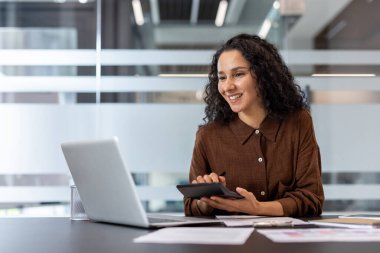 Gülümseyen bir kadın, ofis ortamında dizüstü bilgisayar üzerinde çalışırken hesap makinesi kullanır. Hesapçı gibi görünüyor..