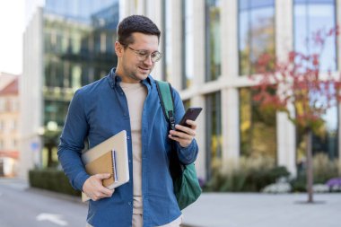 Gözlüklü genç bir adam telefonunu modern bir binanın dışında dizüstü bilgisayar ve defter taşıyor..