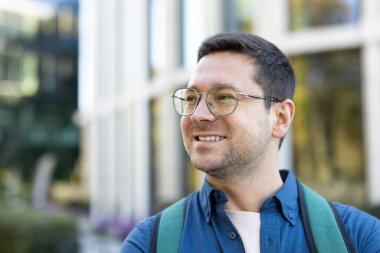 Güneşli bir günde gülümseyen gözlüklü ve sırt çantalı bir adam binanın önünde poz veriyor..