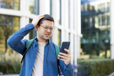 Kafası karışmış bir şekilde telefonuna bakan bir adam, modern pencereli bir binanın yanında kafasına dokunuyor..