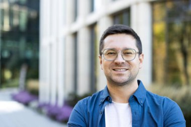 Gözlüklü gülümseyen adam modern bir binanın önünde poz veriyor. Fotoğraf, profesyonel bir ortamda arkadaş canlısı bir kişiyi yakalıyor..