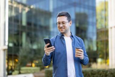Gözlüklü bir adam telefonuna bakıp, modern bir binanın önünde kahve fincanı tutarken gülümsüyor..