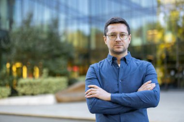 Mavi gömlekli ve gözlüklü bir adam modern bir binanın önünde kollarını kavuşturmuş kameraya bakıyor..