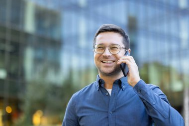 Modern bir binanın önünde gözlüklü, gülümseyen bir adam telefonuyla konuşuyor. İş çağrısı.