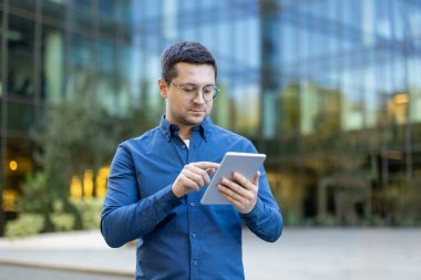 Gözlüklü ve mavi gömlekli bir adam, modern bir binanın önündeki tablete odaklanmış..