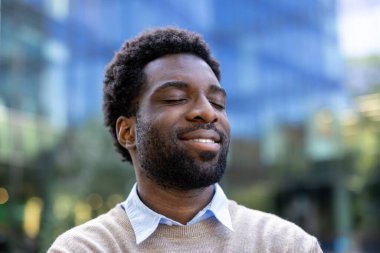 Açık havada gözleri kapalı gülümseyen bir adam. Arkaplanda bulanık bir bina var.