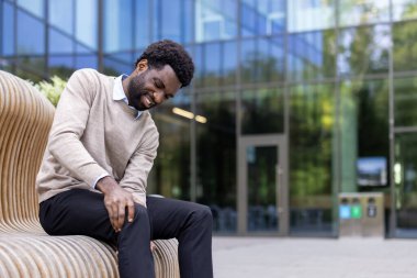 Bir adam dışarıda bankta oturuyor, dizini tutuyor ve acı çekiyor, muhtemelen arka planda modern bir bina var..