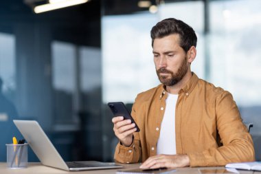 Modern ofis ortamında bir adam, akıllı telefonunu bir masada dizüstü bilgisayar ve diğer iş eşyalarıyla otururken kullanıyor. Işıklandırma yumuşak ve doğal..
