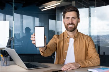 Kahverengi gömlekli gülümseyen bir adam, ofis elemanlarıyla çevrili bir uygulamayı görüntülemek için uygun beyaz ekranlı bir akıllı telefon tutuyor..