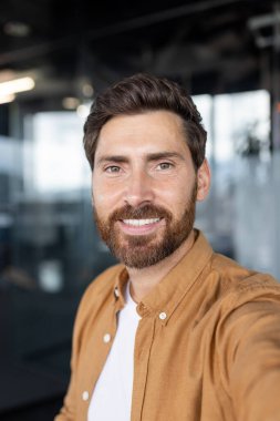 Genç başarılı iş adamı telefon kamerasına bakıyor, video görüşmesi için akıllı telefon uygulamasını kullanıyor, ofis içinde masa başında otururken iş arkadaşları ve arkadaşlarıyla konuşuyor..