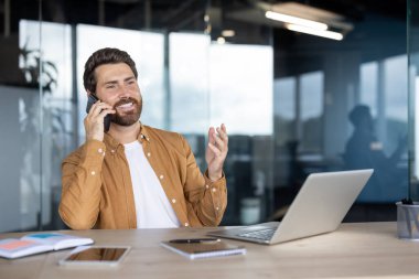 Ofisinde gülümserken telefonla konuşan bir adam. Hoş bir ifadeyle dizüstü bilgisayarıyla çalışıyor..