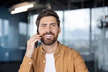 Sakallı, gülümseyen bir adam bir telefon görüşmesi yapıyor. Kapalı alanda, geçmişi bulanık..